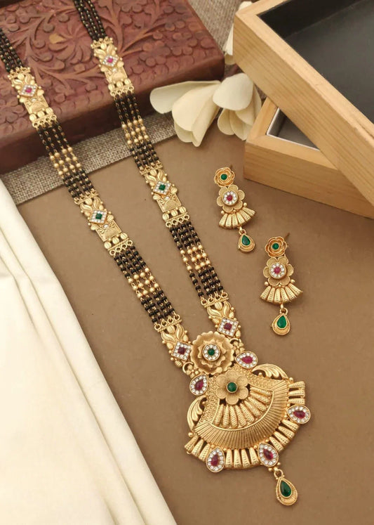 Traditional gold mangalsutra with green and red stones and matching earrings on a beige background